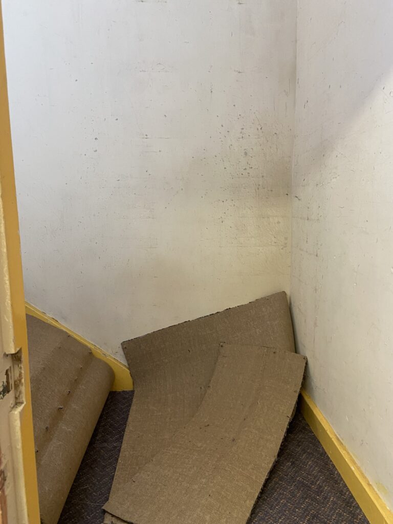 A blank wall and the bottom of a stairwell with some rolled up carpet piled on it
