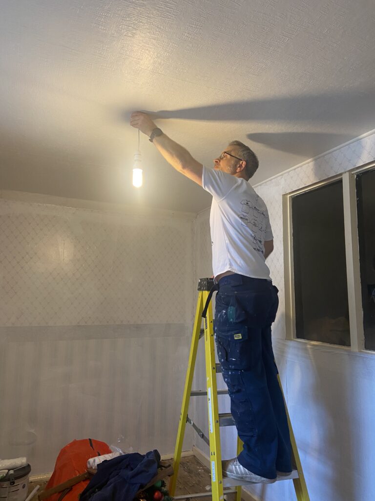 A man on a yellow ladder holding a light fitting against a white ceiling