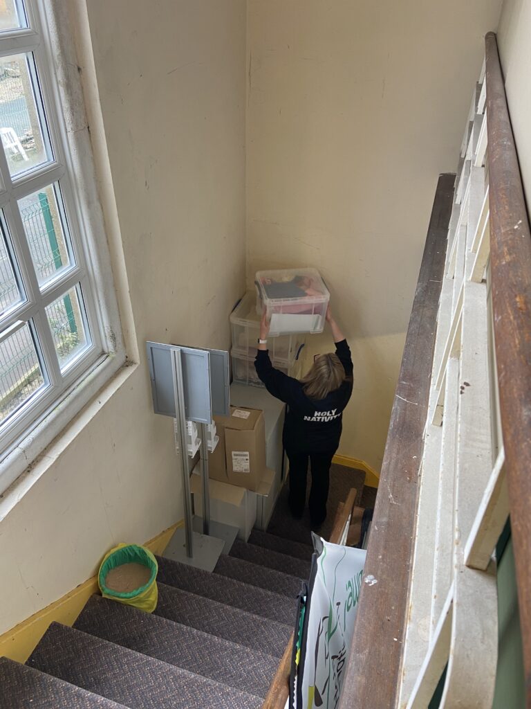 A view from the top of some stairs, a woman stands at the bottom lifting a pile of boxes off the top of a rusty cupboard, there is a bucket of stand and several metal stands on the stairs