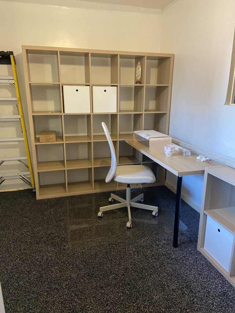 A white painted room with empty shelves, a desk and office chair on a grey carpet