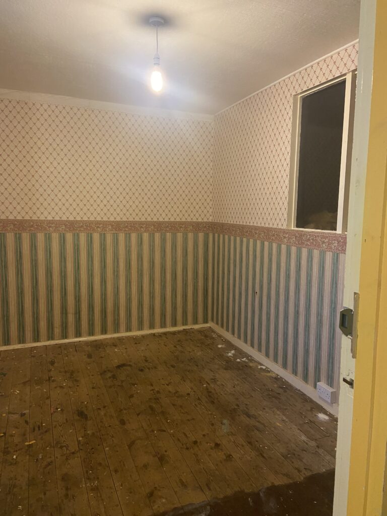 An empty square room with no carpet, there is striped wallpaper on the bottom portion of the wall and diamond patterened wallpaper above