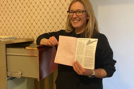 A woman leans against an old metal filing cabinet holding a paper magazine