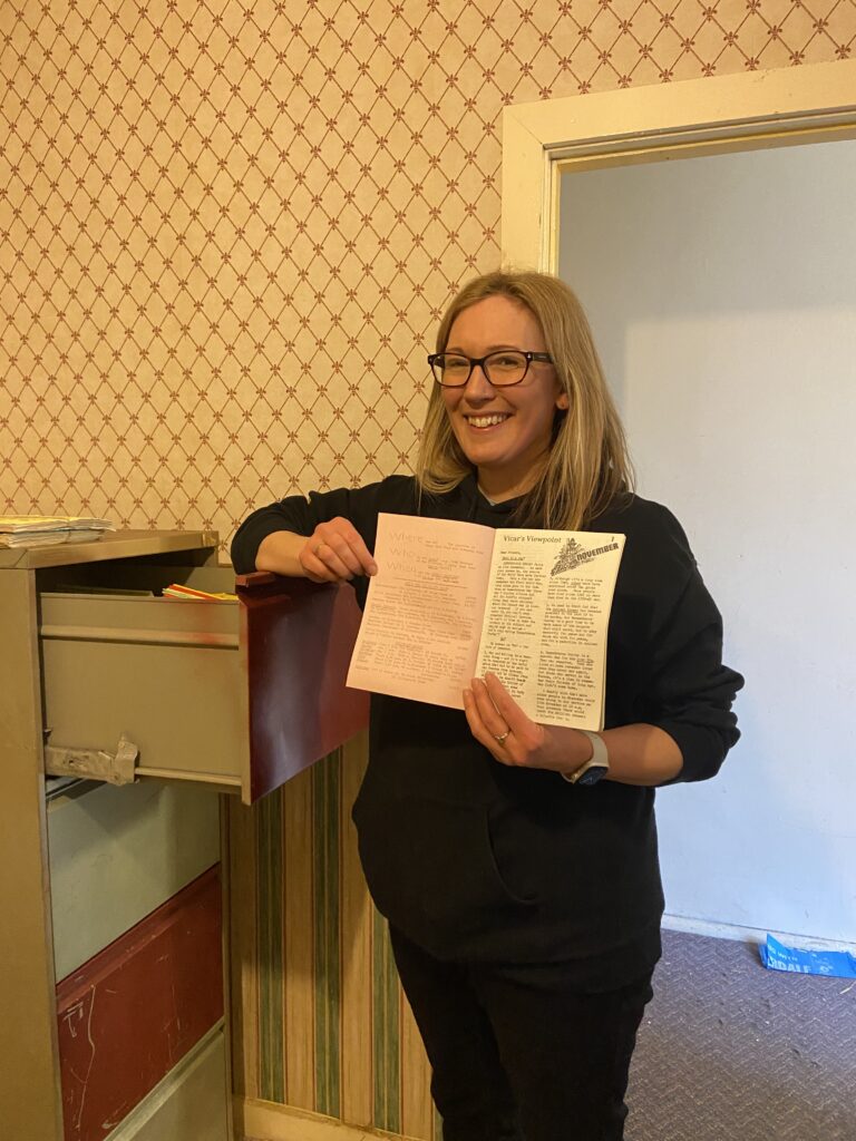 A woman leans against an old metal filing cabinet holding a paper magazine