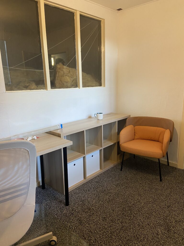 A room with an orange office chair and empty shelves on a grey carpet, there is a dark window behind them and white walls