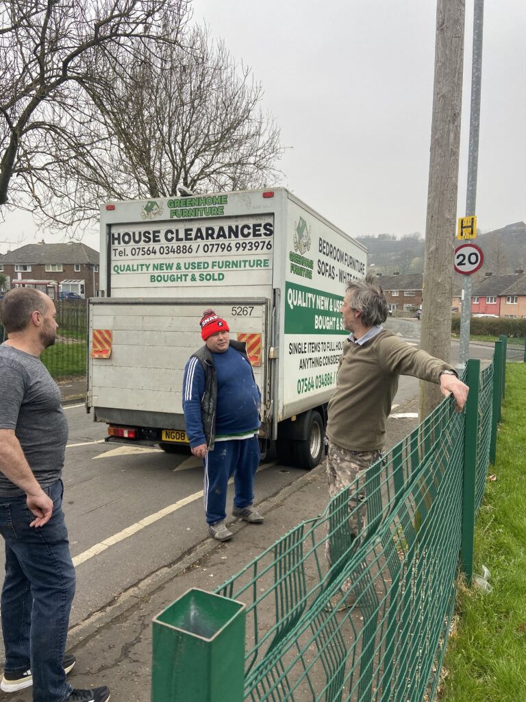 three men standing on the street, one is leaning on a green fence, another is speaking to him wearing a red bobble hat the third is listening. They are standing in front of a white scrap van with green writing on it.