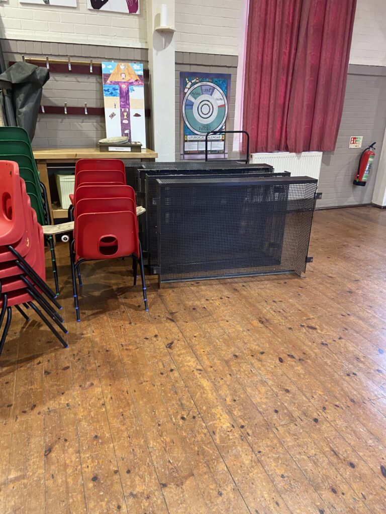 A wooden floored room with stacks of old school chairs in read and some rusty pieces of metal framing