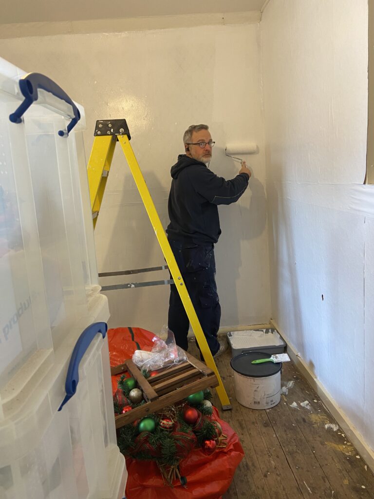 A man in a blue hoody standing behind a yellow ladder holding a paint roller and painting a wall white, he is looking at the camera and smiling