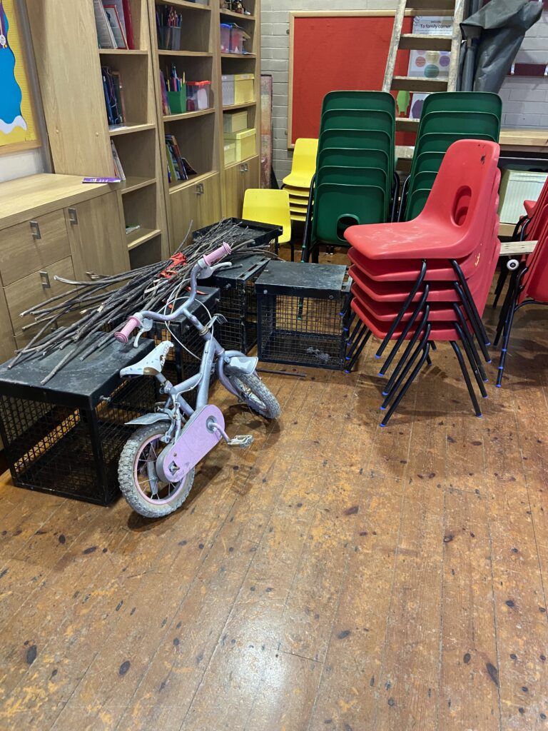 A pile of old school chairs, an old childs bike and rusty metal cages all on a wooden floor leaning against some cupboards.