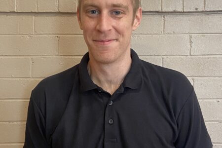 Man with short hair wearing a black poloneck standing in front of a blank wall