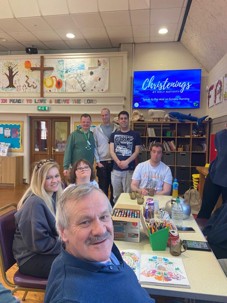 A group of people sitting around a table of activities with others standing in the background. A screen on the wall advertises Christenings.